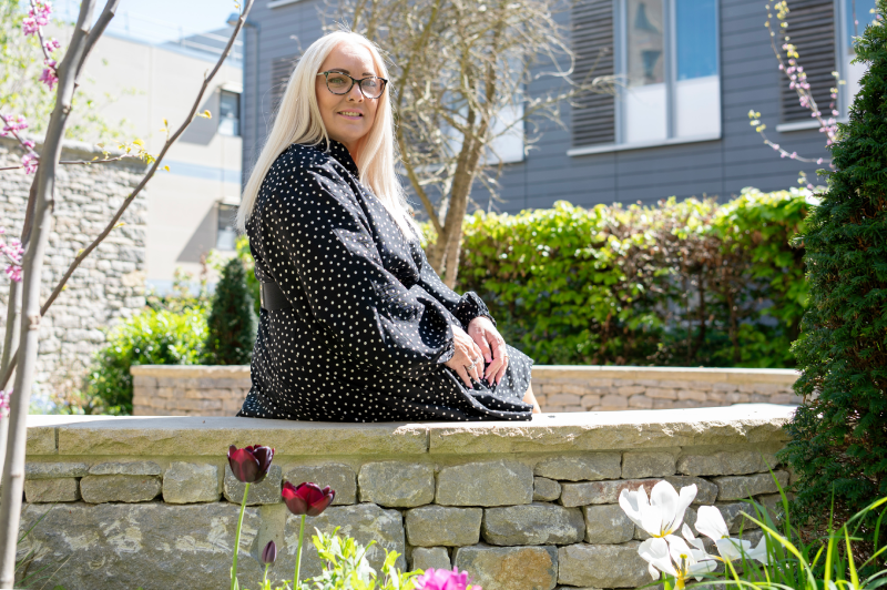 Victoria in the background sitting on a low stone wall looking at the camera across a bed of plants and flowers
