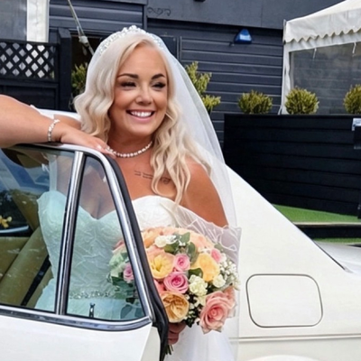 Victoria in a long white dress getting out of a white vintage car holding a floral bouquet in a