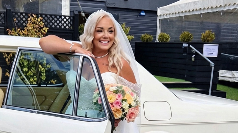 Victoria in a long white dress getting out of a white vintage car holding a floral bouquet in a