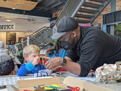 A man with a black mask and cap on helping a young boy create art out of paper at a CUH Arts workshop at Cambridge Junction