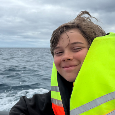 A boy of around 13 in a life jacket on a boat at sea, on a cloudy day with choppy waters. He is smiling