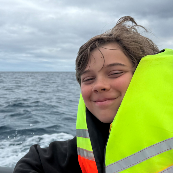 A boy of around 13 in a life jacket on a boat at sea, on a cloudy day with choppy waters. He is smiling