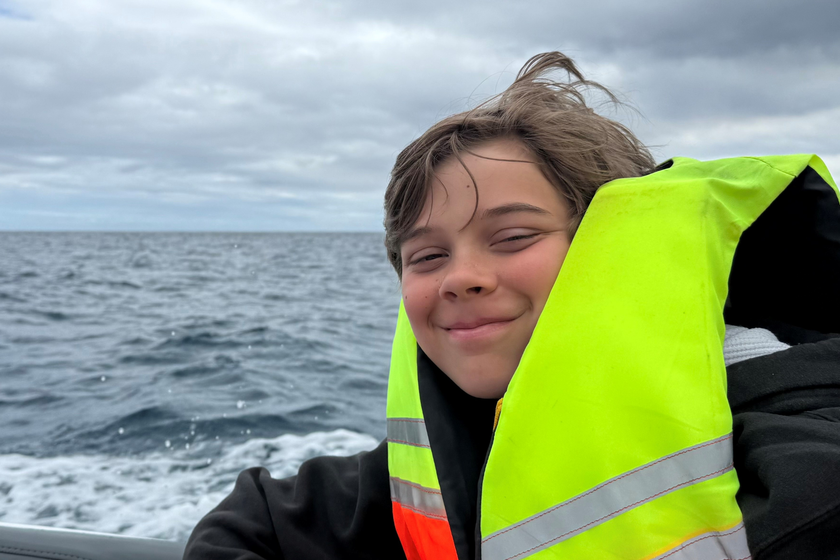 A boy of around 13 in a life jacket on a boat at sea, on a cloudy day with choppy waters. He is smiling