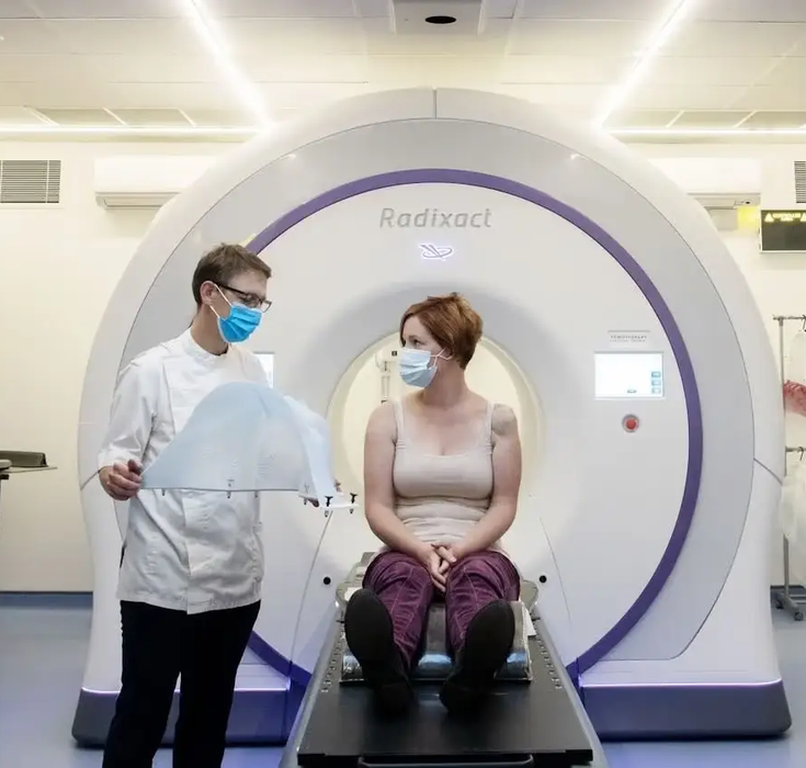 A patient sat on the bed to go into the CT scanner, with a nurse stood next to her holding a radiation guard in his hands