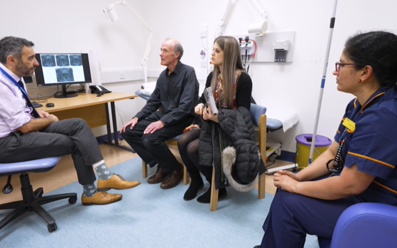 A cancer patient in a consultation room. A doctor and nurse are sat in the room. Images of a scan can be seen on a computer screen. The patient is accompanied by a friend.