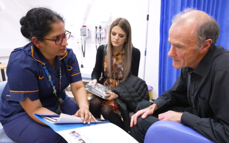 A member of staff showing a cancer patient an information pack. The patient is accompanied by a friend.