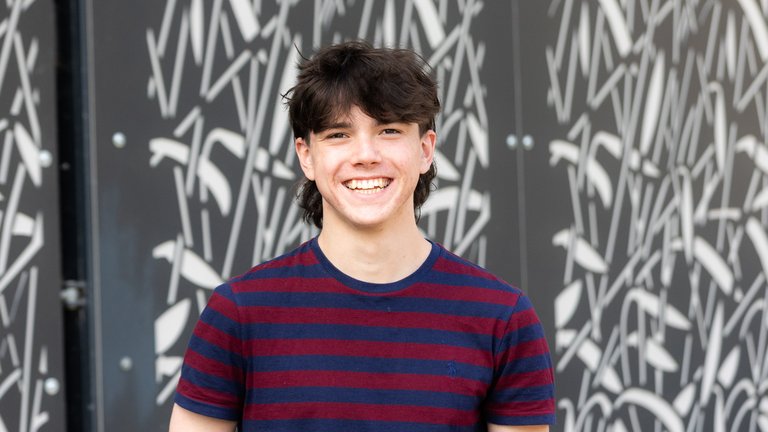 A teenage boy with dark mullet style hair wearing a maroon and navy striped t shirt and jeans, smiling broadly