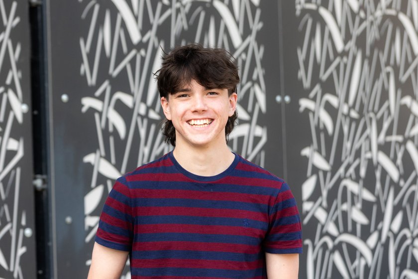 A teenage boy with dark mullet style hair wearing a maroon and navy striped t shirt and jeans, smiling broadly