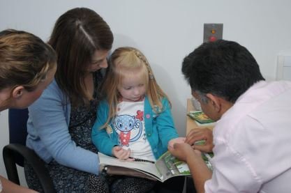A child sitting on her mother's knee reading a book while being seen by a health professional