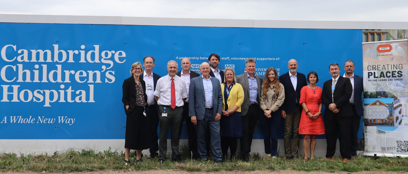 A group of people looking smart and smiling in front of the Cambridge Children's Hospital bright blue hoardings with the logo to the left