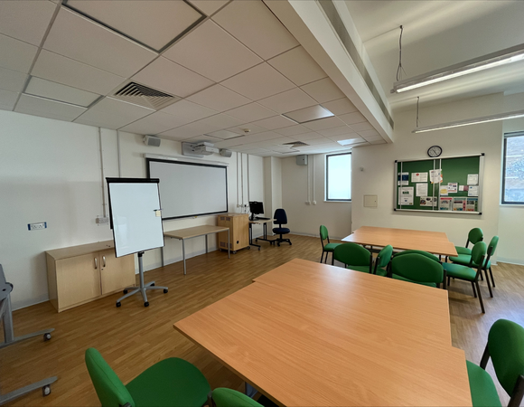 Training room in the Deakin Centre with tables, green chairs, a whiteboard, projector screen, computer desk, noticeboard, and wooden flooring. The room has windows and ceiling lights.