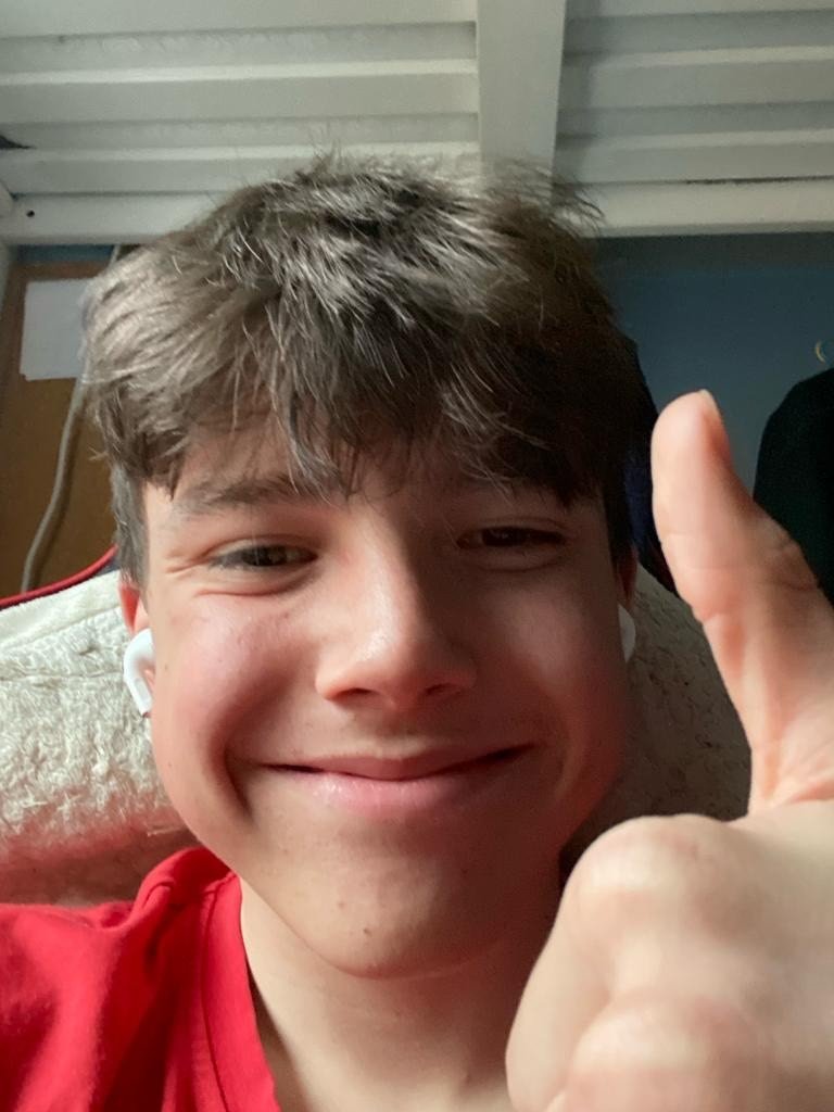 A boy with brown hair and wearing a red polo shirt giving a thumbs up and smiling
