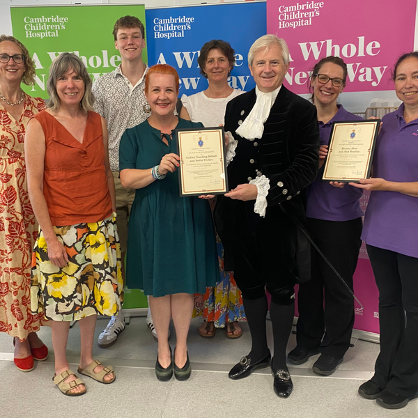 a group of people standing in front of some colourful banners. In the middle holding a certificate out to a woman is a man dressed in a high sheriff outfit, with knickerbockers, a black pirate type hat, a velvet jacket and a ruffled white shirt