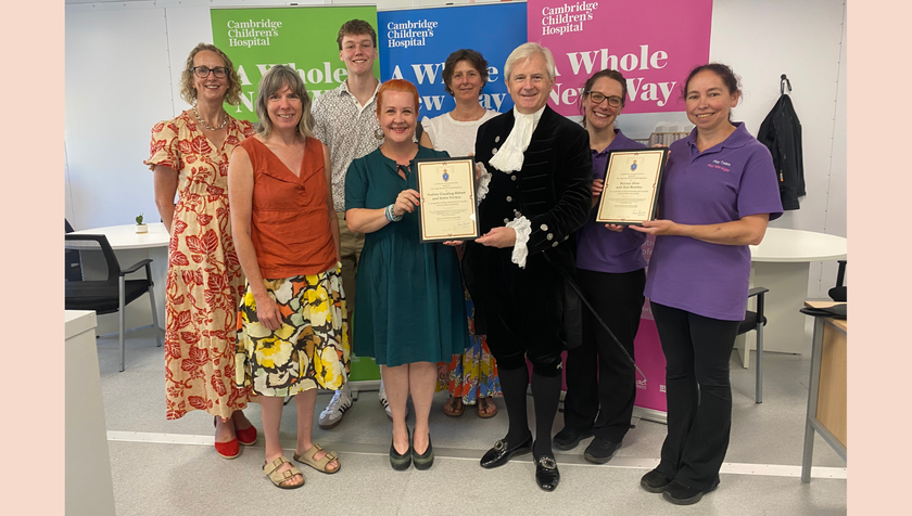 a group of people standing in front of some colourful banners. In the middle holding a certificate out to a woman is a man dressed in a high sheriff outfit, with knickerbockers, a black pirate type hat, a velvet jacket and a ruffled white shirt