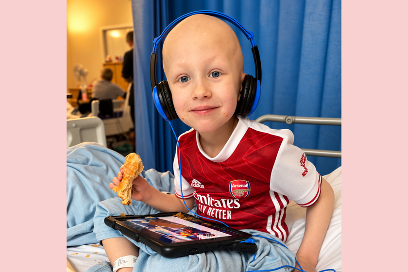 A little boy who has lost his hair through cancer sitting on a hospital bed, cross legged with his ipad on his lap and eating a croissant