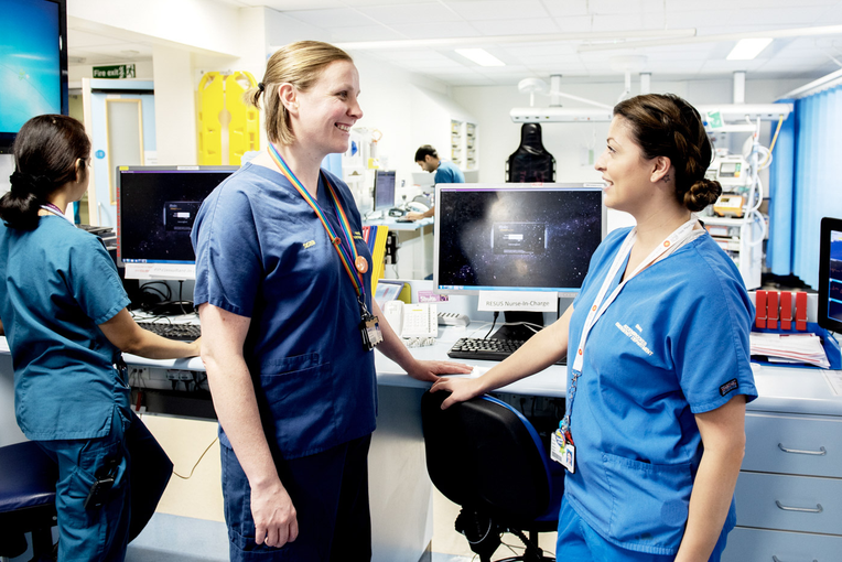 Healthcare staff in a hospital ward stand and work around a nurses' station, with computer monitors, medical equipment and patient bays visible.