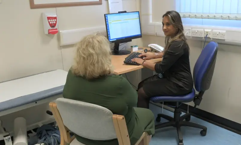 An immunology patient at an appointment, sat with their back to camera, facing a doctor who is at a desk that includes a computer screen and keyboard.