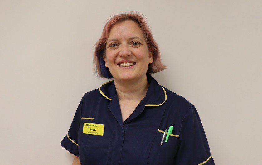Juliette is wearing a dark navy uniform with yellow edging. She has short blond hair, worn in a side parting, with a pale pink streak at the front. She is smiling at the camera. She is wearing a yellow name badge and has two pens in her top pocket.