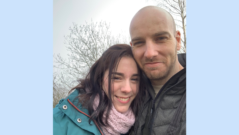 A woman with long dark hair, pink scarf and coat, standing with a tall man with stubbly beard, shaved head, winter coat. Both a smiling