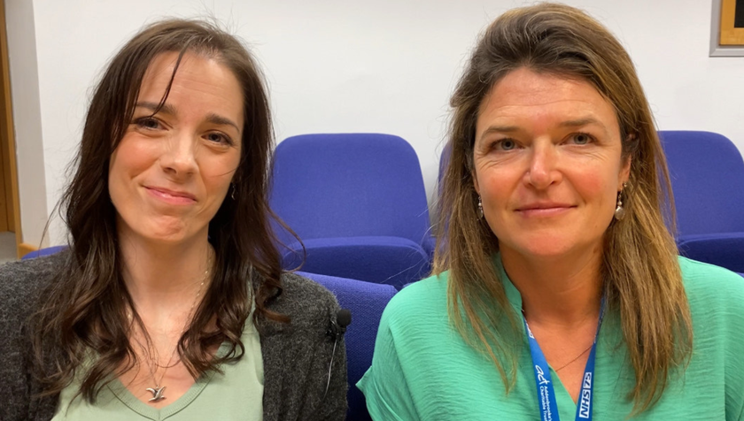 A woman with long dark hair, pale green t shirt and grey cardigan, sitting alongside a woman with long brown hair and a bright green top.