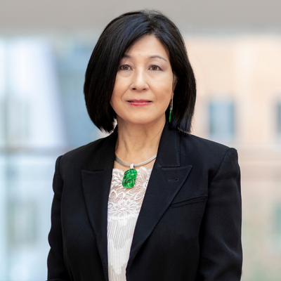 A woman with black glossy hair, a white shirt and black jacket, with a bright green emerald pendant necklace. She is standing in front of a windo