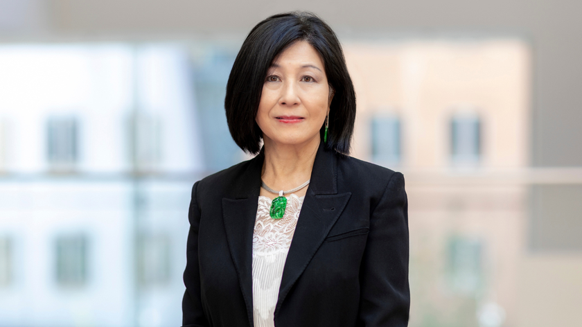 A woman with black glossy hair, a white shirt and black jacket, with a bright green emerald pendant necklace. She is standing in front of a windo