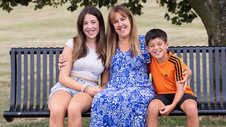 A woman with long hair and a blue and white patterned sun dress, sitting on a park bench with her arms around her children's shoulders. The girl is older with long brown hair. The boy has short dark hair and an orange football top