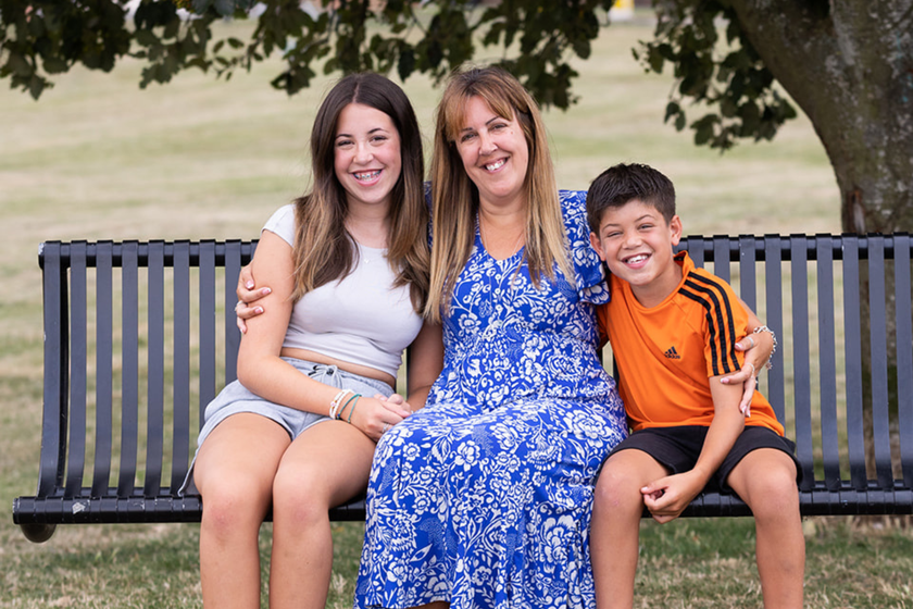 A woman with long hair and a blue and white patterned sun dress, sitting on a park bench with her arms around her children's shoulders. The girl is older with long brown hair. The boy has short dark hair and an orange football top