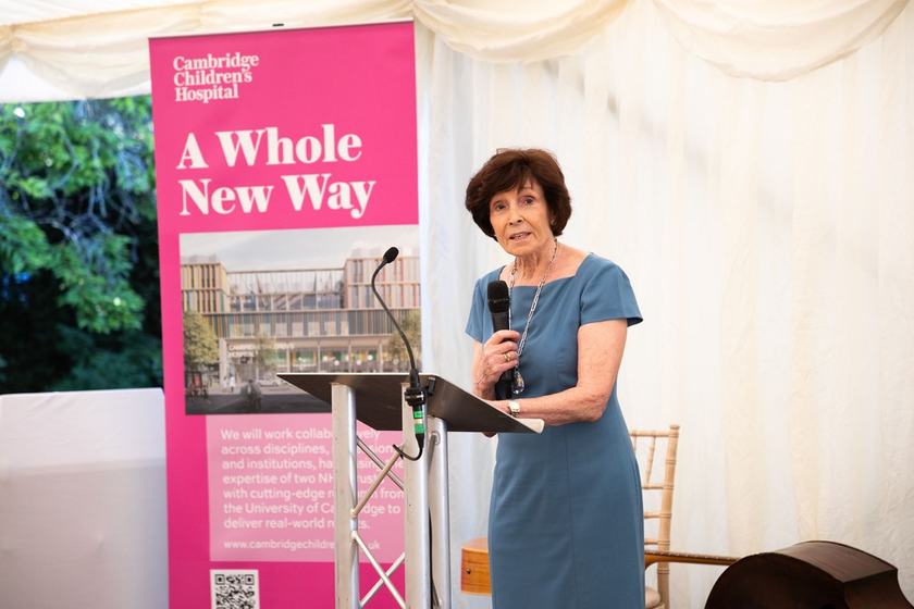 A woman standing at a lecturn on a stage in a marquee giving a speech. She has dark brown short hair and is wearing a denim blue short sleeved dress
