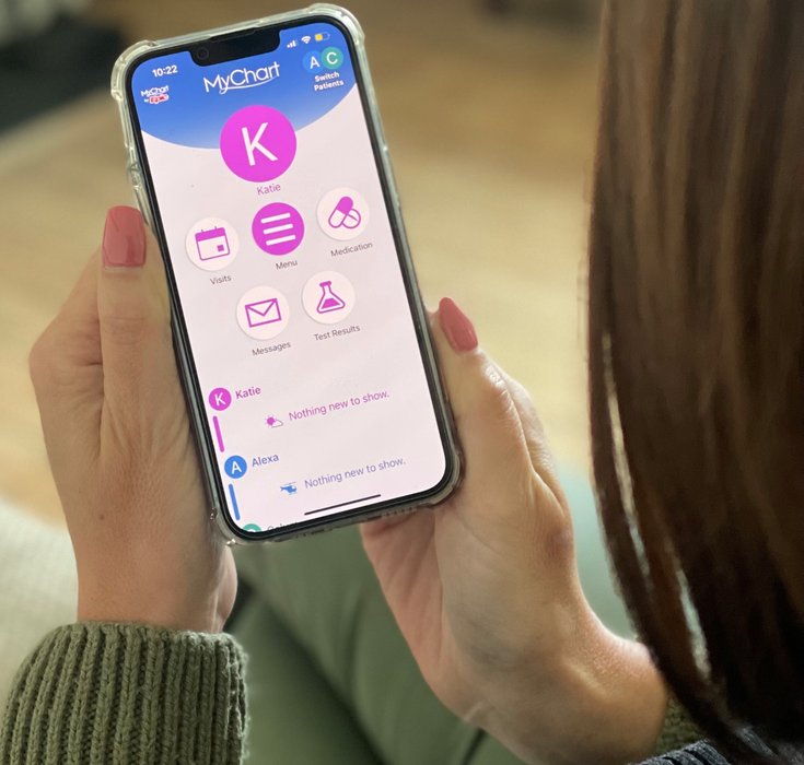 Hands holding a mobile phone displaying the MyChart app. Pink round icons are shown labelled 'visits', 'menu', 'medication', 'messages', 'test results' and a profile icon labelled 'Katie' at the top.
