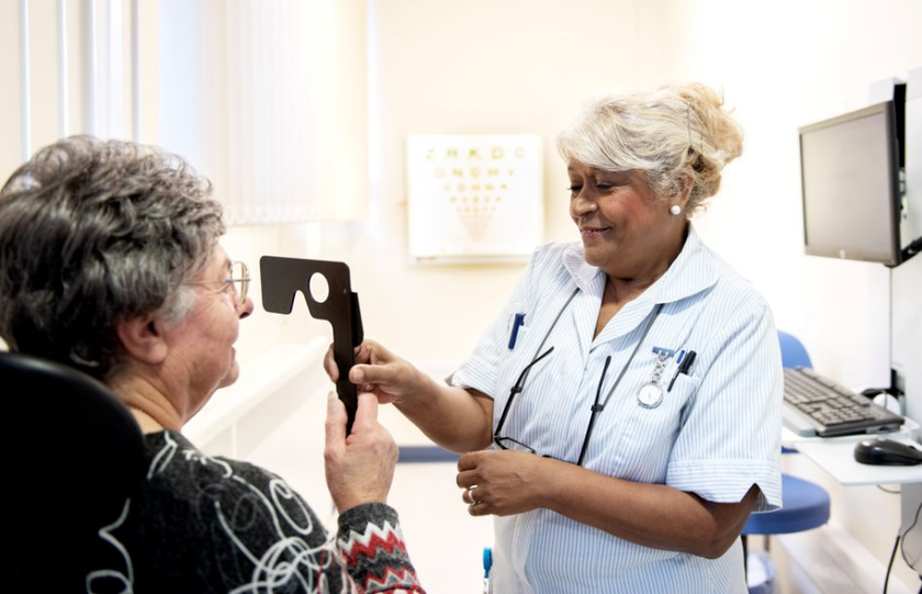 Patient undergoing a vision assessment in a clinic, holding an eye test occluder while a healthcare worker provides support.