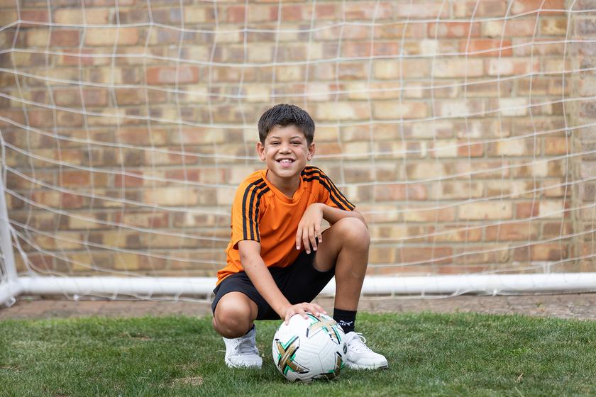 A young boy wearing an orange football top and black shorts. He is crouching down in front of a football goal with a football at his feet
