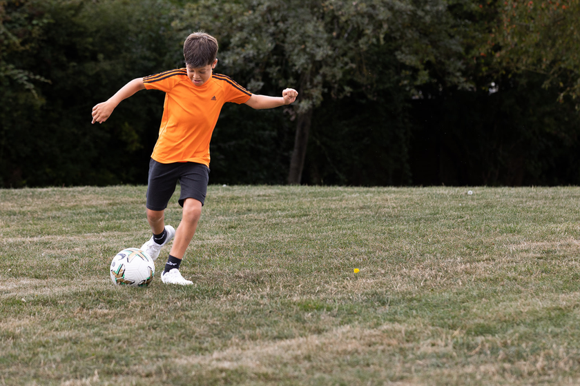 A boy with short dark hair and an orange football top kicking a football in the park