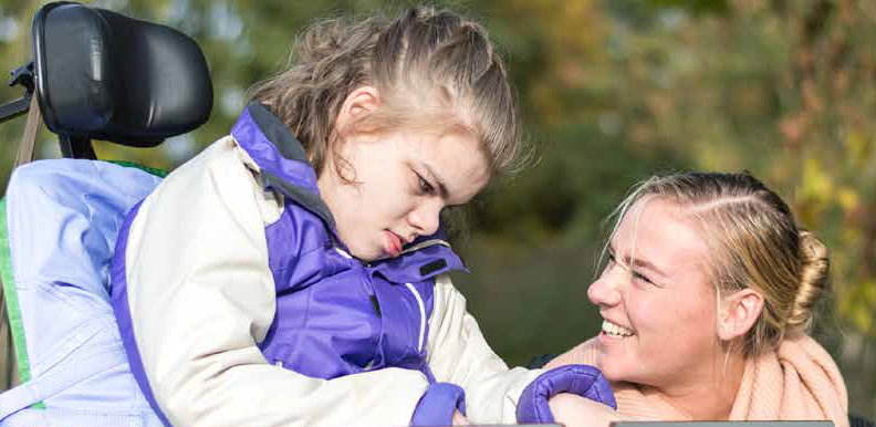 A young person in a wheelchair with their carer. The carer is smiling and there is a blurred background of showing trees