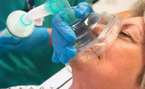 A patient being administered general anaesthetic. Patients laying down with eyes closed a mask and tube can be seen placed over the patients mouth and nose by an anaesthetist wearing blue gloves.