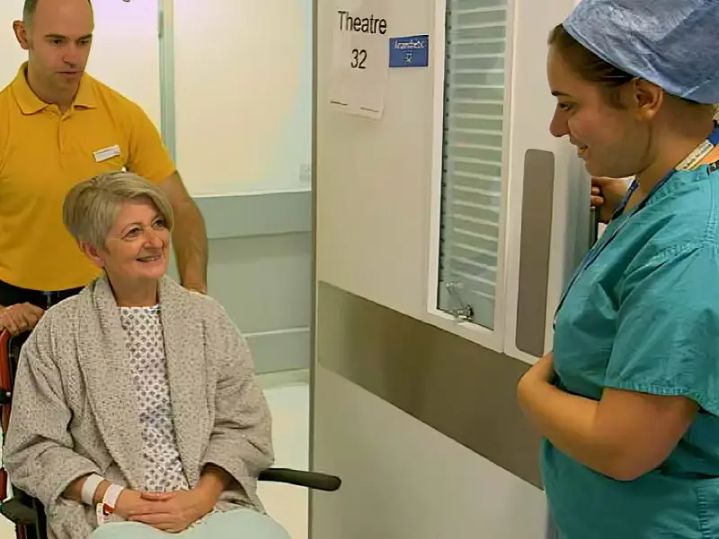A patient wearing a theatre gown, dressing gown and blanket over knees is sat in a wheelchair. A porter in a yellow polo shirt is stood behind. The patient is met by a member of staff in scrubs, standing in the doorway at the entrance to theatre.