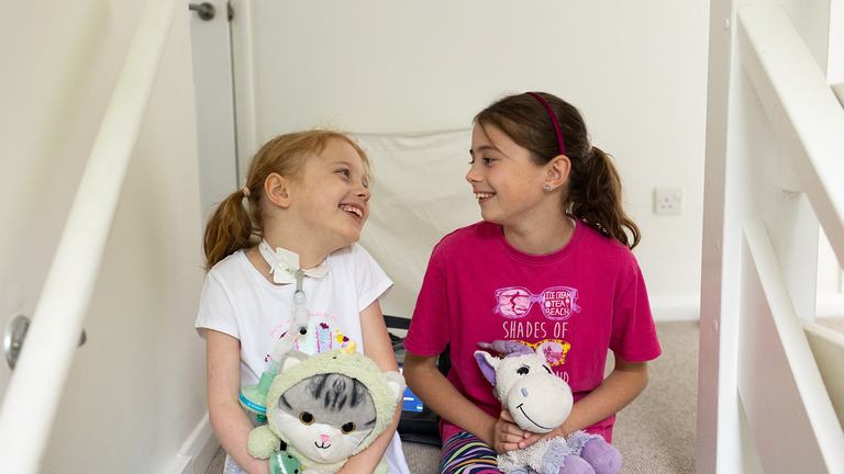 Two girls sitting on the stairs. One had gingery hair and is holding a cat teddy. The other has long brown hair and is holding a unicorn teddy. They are looking at each other and laughing