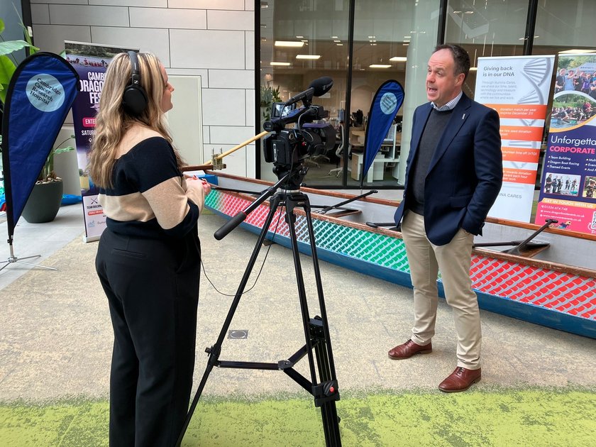 A man in a suit talking to a TV camera and journalist, standing in front of a dragon boat