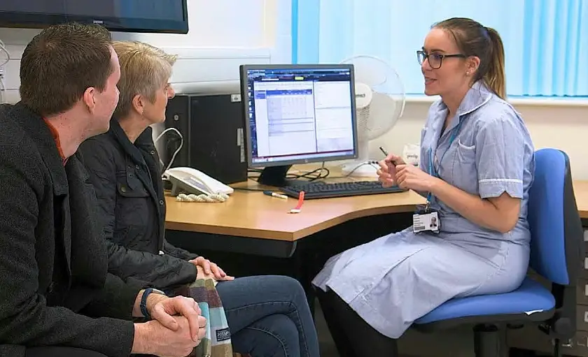 A patient and loved one sat talking to a nurse who is sat opposite at a desk.