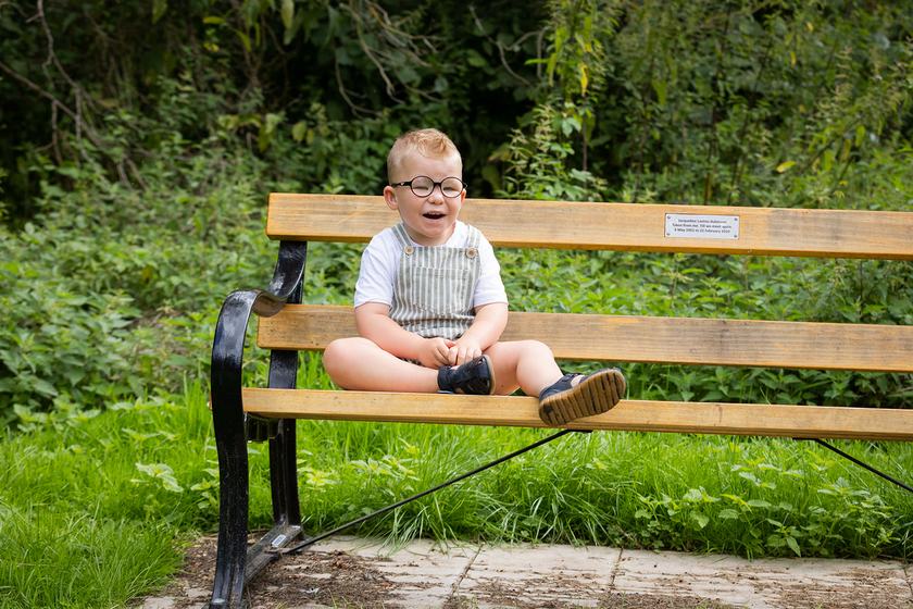 A little boy with blond short hair and round rimmed glasses sitting on a wooden bench outside in the sunshine. He is wearing dungaree shorts and a white t shirt