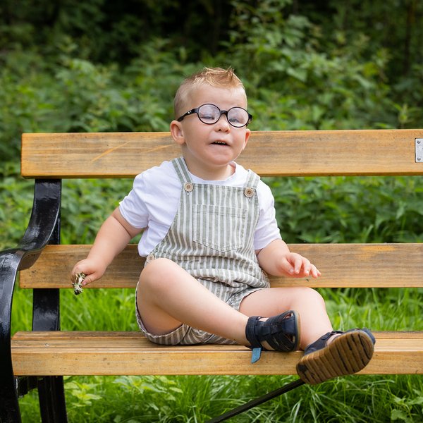 A little boy with round glasses sitting on a bench outside. He's wearing green and white stripy dungaree shorts, black shoes and a white t shirt