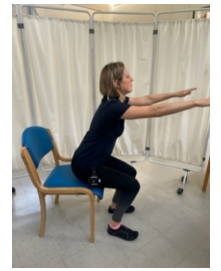 A woman sat on the edge of a chair with her feet apart on the ground and her arms stretched out in front of her while leaning slightly forward