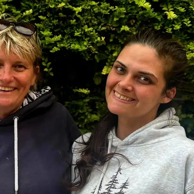 Vanessa and Nicole side by side smiling into the camera with an arm around each other. Standing outside in front of a large green hedge.