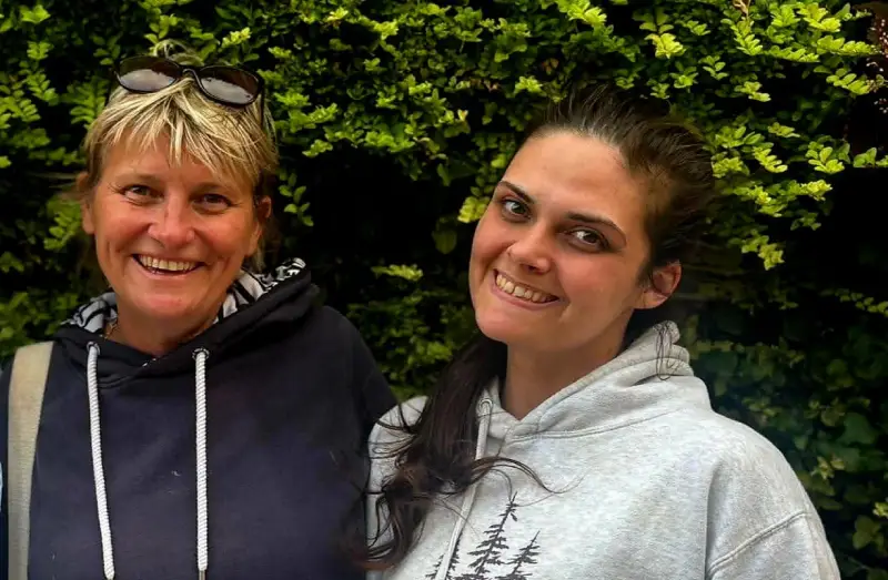 Vanessa and Nicole side by side smiling into the camera with an arm around each other. Standing outside in front of a large green hedge.