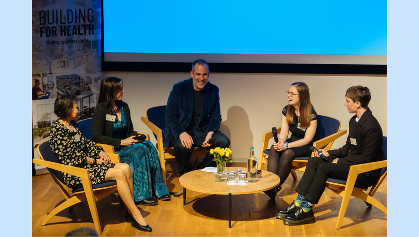 A man with dark grey hair and dark grey stubble, in a suit, on the stage with a group of young adults and parents. They are having a discussion at a conference